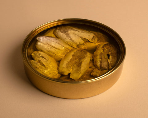 An open tin of hake cheeks in golden coloured olive oil, in a round golden coloured tin, against a pale background.