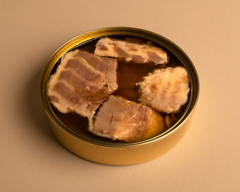 An open tin of sea bass fillets in a round, golden round on a pale background.