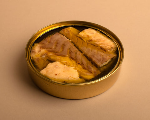 A round golden tin with fillets of sea bass in olive oil, set against a pale background.