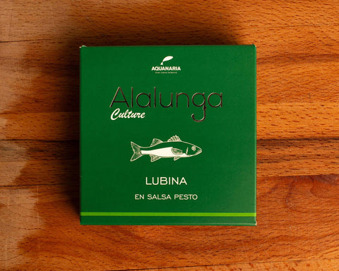 A tin of sea bass in pesto in green card packaging against a wood background. The packaging has Alalunga written in silver lettering above a white illustration of a sea bass, and a green strip along the bottom.