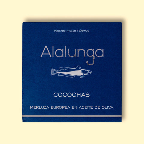 A tin of hake cheeks in olive oil in square-shaped blue card packaging against a yellow table background. The packaging has Alalunga written in silver lettering above a white illustration of a hake.