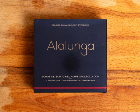 Canned white tuna in square blue card packaging with Alalunga in silver, embossed lettering above a diagram of a tuna. There is a purple stripe along the bottom of the packaging.