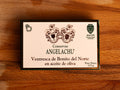 A tin of ventresca or tuna belly in a rectangular card packaging. The box is white with a dark green and golden border. Conservas Angelachu is written in dark green lettering and ventresca de Bonito del Norte en aceite de oliva in gold leaf lettering, beneath a coat of arms. The box is against a wood background.