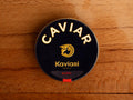 A circular dark blue tin of caviar on a wood backdrop. Caviar is written in white lettering at the top of the tin, above the Kaviari logo: an illustration in gold of a sturgeon fish above Kaviari Paris also in gold lettering. The tin is sealed with a red and blue label reading Baeri Royal.