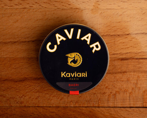 A circular dark blue tin of caviar on a wood backdrop. Caviar is written in white lettering at the top of the tin, above the Kaviari logo: an illustration in gold of a sturgeon fish above Kaviari Paris also in gold lettering. The tin is sealed with a red and blue label reading Baeri Royal.