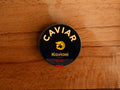 A circular dark blue tin of caviar on a wood backdrop. Caviar is written in white lettering at the top of the tin, above the Kaviari logo: an illustration in gold of a sturgeon fish above Kaviari Paris also in gold lettering. The tin is sealed with a red and blue label reading Baeri Royal.