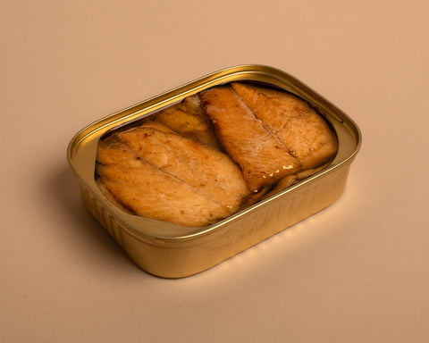 An open rectangular golden tin of herring in olive oil and lemon extract, set against a pale background.