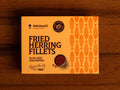 A tin of herring fillets in pale orange packaging, against a wood background. There is a small bowl of pink peppercorns on the packaging.