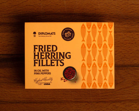A tin of herring fillets in pale orange packaging, against a wood background. There is a small bowl of pink peppercorns on the packaging.