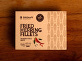 A tin of herring in pale brown card packaging with brown lettering by a picture of two whole red chillies next to two garlic cloves and two slices of red chilli pepper.