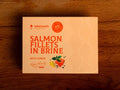 A tin of salmon fillets in pale orange packaging with orange lettering by a picture of two salmon fillets and two lemon wedges with a sprig of basil.