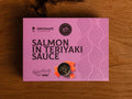 A can of salmon in purple card packaging with dark purple lettering. There is an image of diced salmon by a bowl of teriyaki sauce.