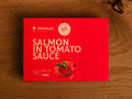 A tin of salmon in red card packaging with pale grey lettering above an image of a diced salmon fillet round a bowl of tomato sauce topped with a sprig of basil.