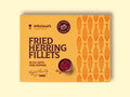 A tin of herring fillets in pale orange packaging, against a wood background. There is a small bowl of pink peppercorns on the packaging.