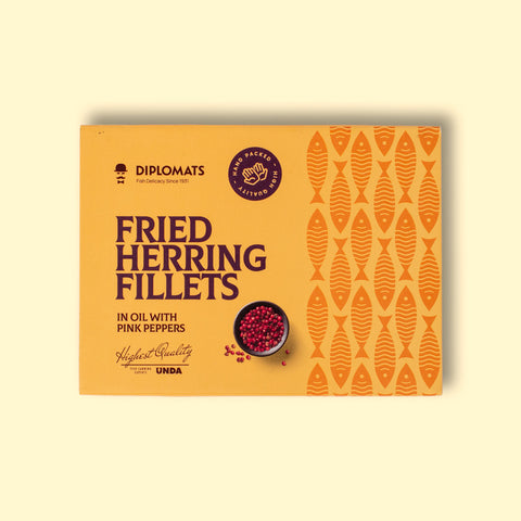 A tin of herring fillets in pale orange packaging, against a wood background. There is a small bowl of pink peppercorns on the packaging.