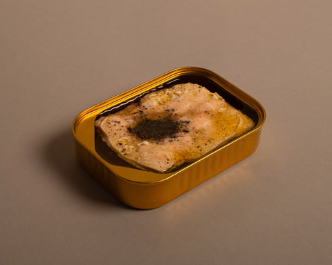 An open golden tin of pink salmon fillet topped with a sprig of lemon verbena herb, set against a pale background.