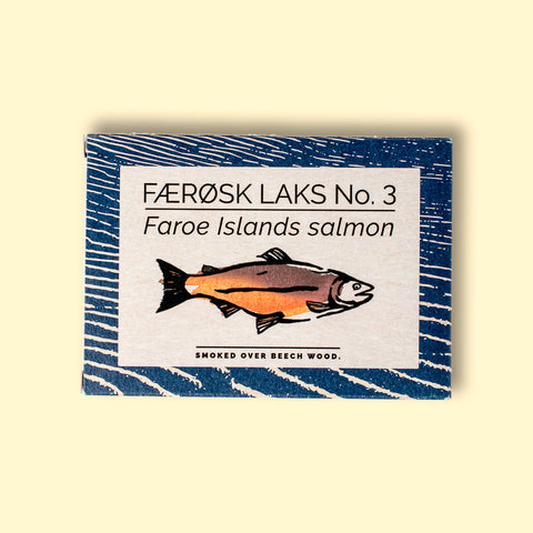 Tinned Faroe Islands Salmon with white and blue packaging, featuring an illustration of a salmon with text beneath reading smoked over beech wood.