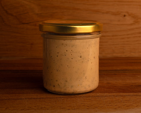 A glass jar of mustard with dill and a golden cap, placed against a wooden background. The jar is transparent, showing the mustard inside.