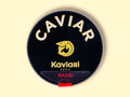 A circular dark blue tin of caviar on a wood backdrop. Caviar is written in white lettering at the top of the tin, above the Kaviari logo: an illustration in gold of a sturgeon fish above Kaviari Paris also in gold lettering. The tin is sealed with a red and blue label reading Baeri Royal.