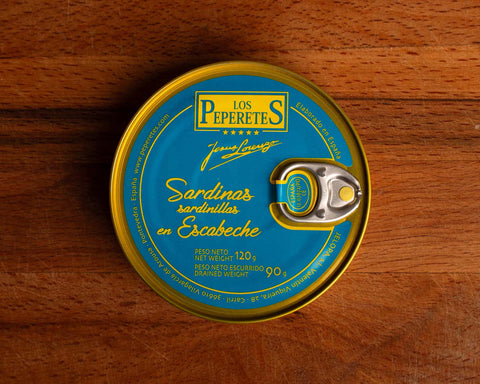 A tin of sardines in escabeche, featuring light blue packaging with golden corners and the Los Peperetes label at the top, set against a wooden backdrop