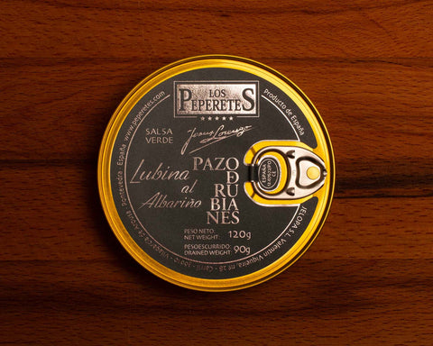 Sea bass in salsa verde with albarino wine, packaged in a circular black tin with golden corners and silver lettering, against a wooden backdrop.