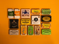Collection of 15 tins on an orange background. The tins are colourful, some of them have vintage illustrations, some of them are British, some are Spanish.