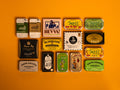 Collection of 15 tins on an orange background. The tins are colourful, some of them have vintage illustrations, some of them are British, some are Spanish,