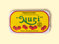A tin of sardines in a yellow paper wrap with the Nuri logo in green on the front above some images of tomatoes. The tin has a red and white border and is on a wood backdrop.