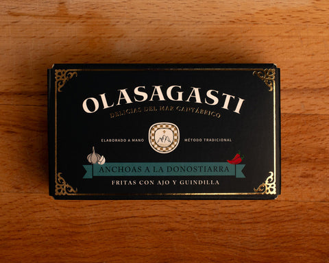 A tin in rectangular, dark blue card packaging, with an ornate gold-coloured border and Olasagasti in white lettering across the top above an illustration of a green banner with Anchoas a la Donostiarra written on it and illustrations of red chillies and garlic above it.