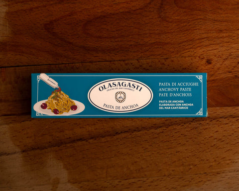 Anchovy paste in a long, rectangular blue and white card box against a wood background. The box has an illustration of a dish of spaghetti being topped with the anchovy paste next to the Olasagasti logo.