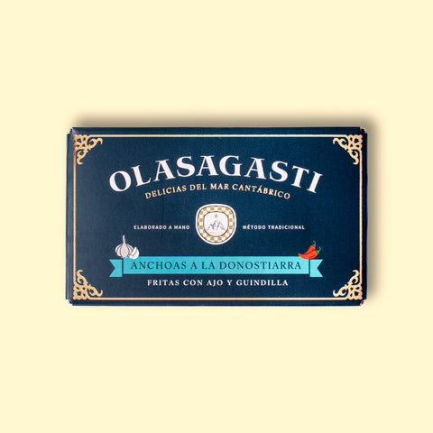 A tin in rectangular, dark blue card packaging, with an ornate gold-coloured border and Olasagasti in white lettering across the top above an illustration of a green banner with Anchoas a la Donostiarra written on it and illustrations of red chillies and garlic above it.