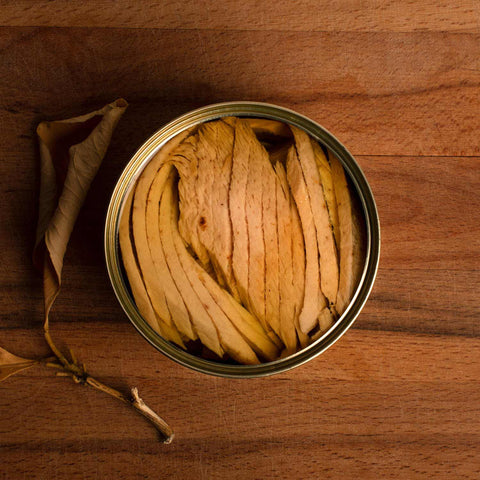 An open circular tin of ventresca fillet layers, placed against a wooden background with a dry brown leaf beside it.