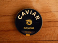 A circular dark blue tin of caviar on a wood backdrop. Caviar is written in white lettering at the top of the tin, above the Kaviari logo: an illustration in gold of a sturgeon fish above Kaviari Paris also in gold lettering. The tin is sealed with a yellow and blue label reading Osciètre prestige.