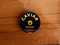 A circular dark blue tin of caviar on a wood backdrop. Caviar is written in white lettering at the top of the tin, above the Kaviari logo: an illustration in gold of a sturgeon fish above Kaviari Paris also in gold lettering. The tin is sealed with a yellow and blue label reading Osciètre prestige.