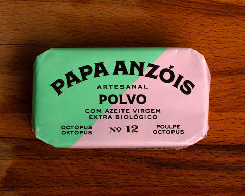 A rectangular tin of octopus in extra virgin olive oil with green and pale lilac paper packaging against a wood background.