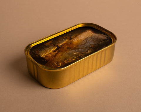 A tin of sardines in olive oil inside an open golden can, with two silvery coloured fish in golden olive oil visible.