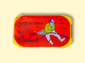 A tin of sardines in a red paper wrap with a yellow border. A fisherman in a green jersey mends his net on the front. The tin is on a wood background.