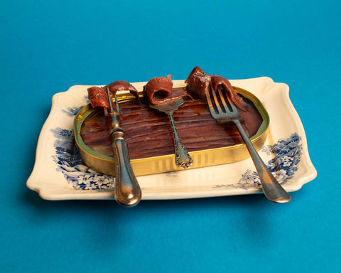 An open golden tin of anchovies placed on a plate, with three different forks each holding a piece of anchovy, set against a blue backdrop.
