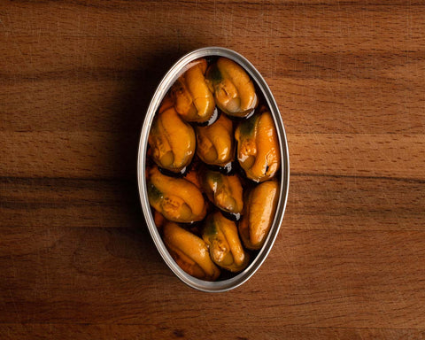 An open silver tin of mussels in rapeseed oil, against a wooden backdrop.