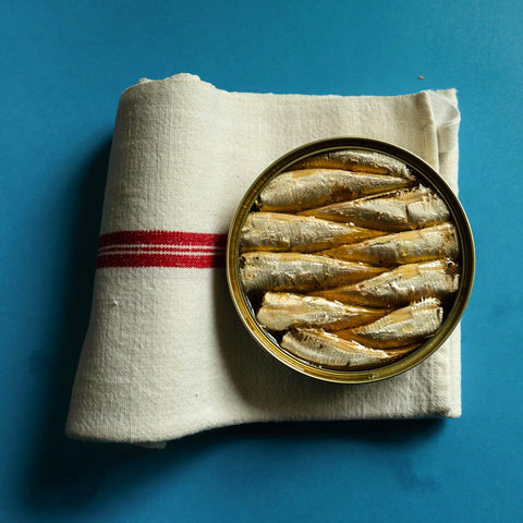 An open golden tin of sardines placed on a white tablecloth, set against a blue backdrop.