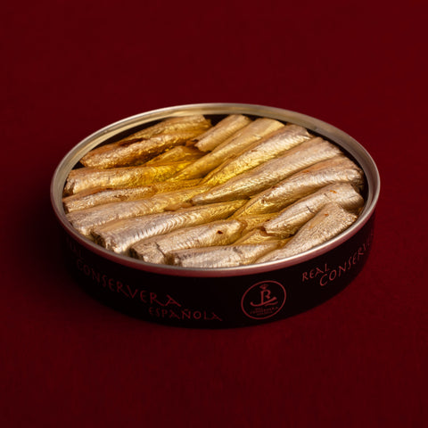 A black circular tin with silver borders containing small, bright silver sardines, featuring the Real Conservera logo, set against a dark red background.