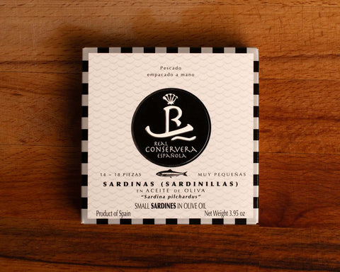A tin of small sardines in black and white card packaging with a black and silver chequered border, against a wood background.