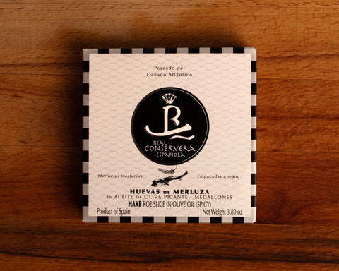 A tin of hake roe slices in olive oil in white packaging, with a silver and black chequered border, featuring an illustration of a hake fish and chilli, against a wooden backdrop.