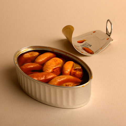 seven bright orange mussels in transparent pickled sauce, inside an oval tin with the peeled tin cover resting behind, set against a pale backdrop.