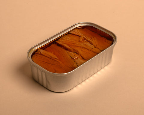 A tin of tuna fillets in red pepper infused olive oil, packaged in a silver tin set against a pale background.