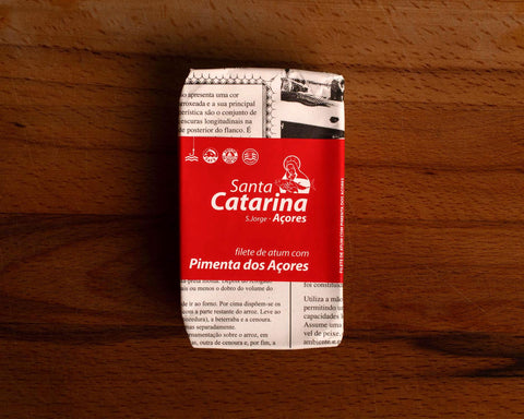 A tin of tuna with pepper, in newspaper packaging with a red band around it with Santa Catarina written in white lettering on it.