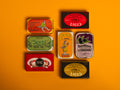 A selection of colourfully-packaged tinned scallops, tuna, mackerel and sardines against a yellow background.