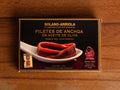 Anchovy fillets in rectangular packaging with brown borders, featuring an image of filleted anchovies resting on a spoon.