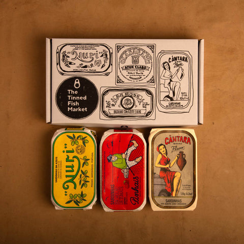 Three colourfully packaged tins of fish beneath a gift box illustrated with line drawings of the tins and The Tinned Fish Market logo. Against a light brown background.