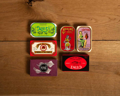 Six tins of fish in brightly coloured packaging  against a wood background.